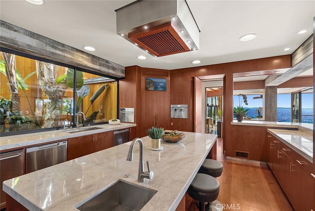 This modern kitchen features warm wood cabinetry and a large central island topped with a light-toned stone surface. A prominent stainless steel range hood hangs above the island, while large windows provide a view of lush greenery on one side and a scenic ocean vista on the other. The space is designed with a clean, contemporary aesthetic that emphasizes natural light and seamless indoor-outdoor integration.