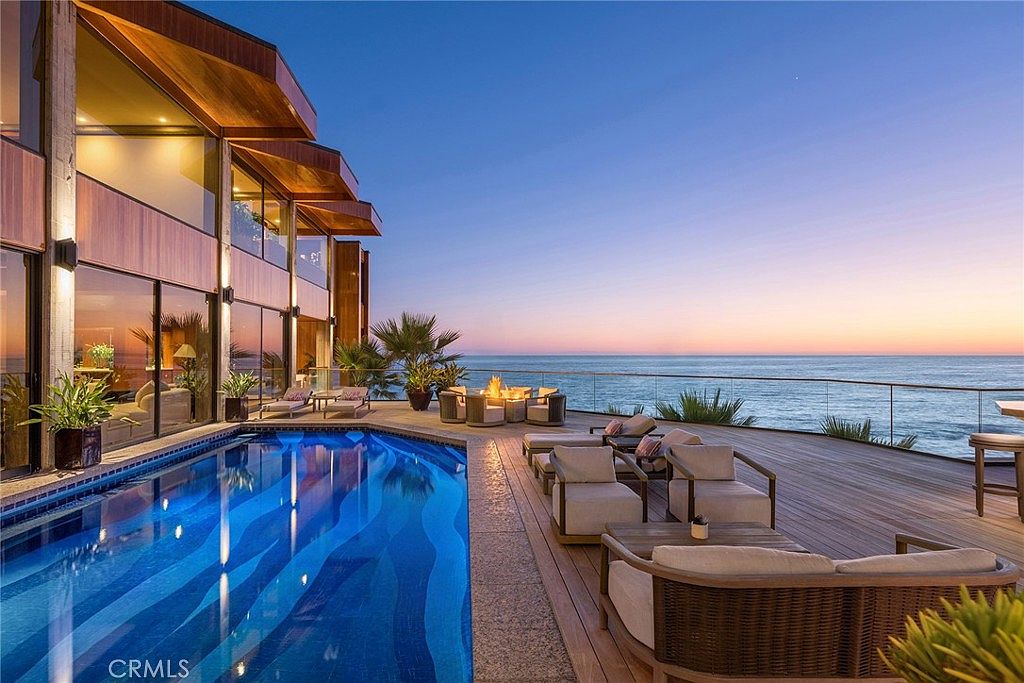 This stunning oceanfront terrace features a sleek, rectangular swimming pool with a vibrant blue tiled interior that reflects the sunset sky. A spacious wooden deck surrounds the pool, furnished with comfortable lounge chairs and a central fire pit area, all overlooking the expansive Pacific Ocean. The modern residence showcases extensive glass walls that provide a seamless transition between the indoor living spaces and the outdoor entertainment area.