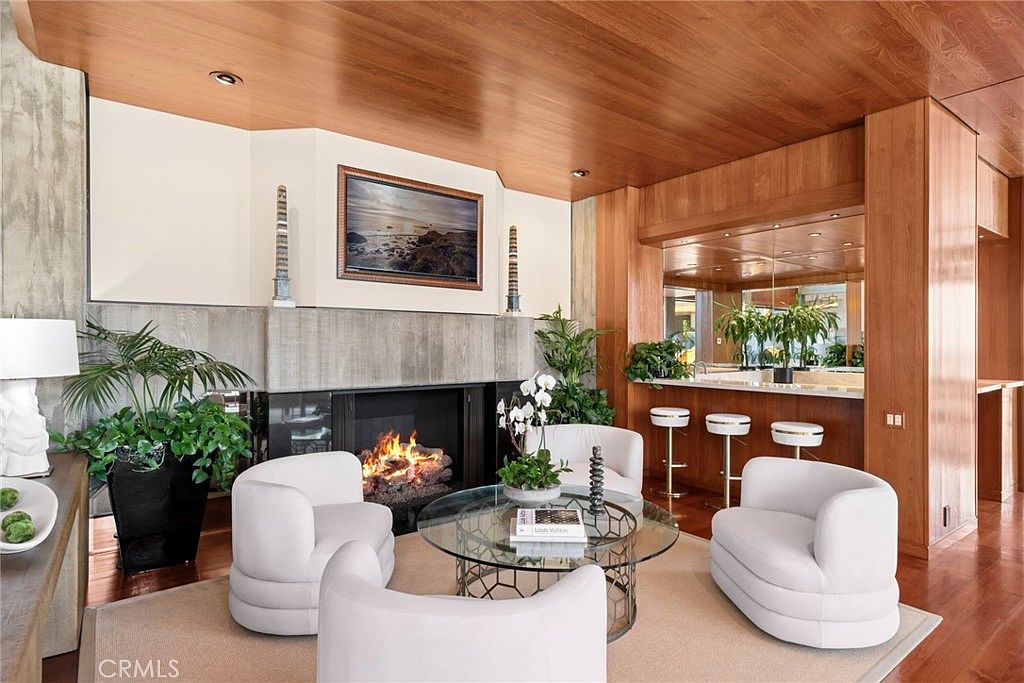 This sophisticated living room features a warm, wood-paneled ceiling and a prominent stone-clad chimney breast surrounding a fireplace. The space is furnished with three plush, cream-colored curved chairs arranged around a circular glass coffee table, set atop a neutral area rug. To the right, a built-in bar area with stools and mirrored backing adds a luxurious touch, while large potted plants provide a vibrant contrast to the rich wood tones.
