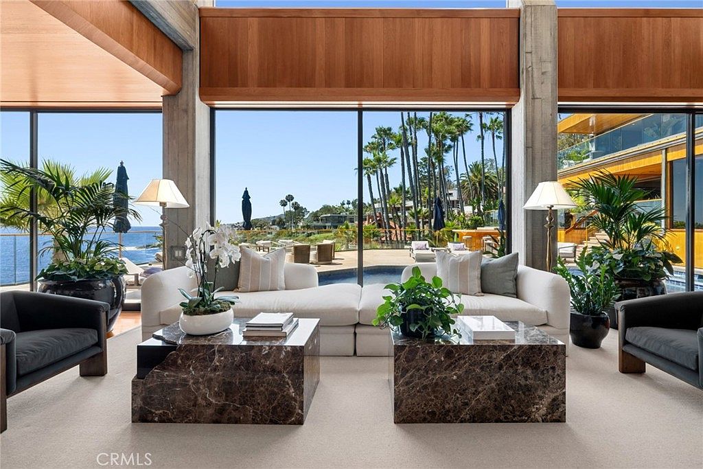 This sophisticated living room features a symmetrical layout with two plush white sofas facing each other across a pair of dark, polished stone coffee tables. Large floor-to-ceiling windows provide an expansive, unobstructed view of the ocean and a private pool area, bathing the space in natural light. The room is accented by tall indoor plants, elegant table lamps, and warm wood paneling above the windows, creating a luxurious and serene atmosphere.
