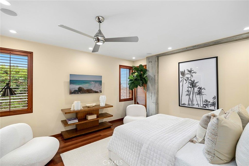 This bright and airy bedroom features a neutral color palette with warm wood flooring and a textured white bedspread. A modern ceiling fan centers the room, while a console table with a coastal-themed art piece and a large palm-tree print on the wall add a relaxed, sophisticated aesthetic. The space is furnished with two plush, rounded accent chairs, creating a comfortable and inviting atmosphere.
