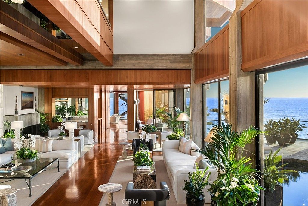 This expansive, open-concept living area features rich wood paneling and warm wood flooring, creating a sophisticated and inviting atmosphere. The space is defined by large glass walls that offer panoramic ocean views, seamlessly blending indoor living with the coastal landscape. Furnished with plush white sofas and contemporary decor, the room is illuminated by natural light and elegant lamps, emphasizing a luxurious, airy aesthetic.