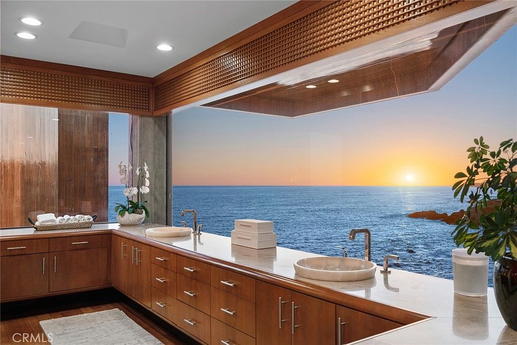 This luxurious primary bathroom features a long, L-shaped vanity with warm-toned wood cabinetry and a light-colored stone countertop. The space is defined by expansive, wall-to-wall glazing that provides an unobstructed, panoramic view of the ocean at sunset. The minimalist design emphasizes the connection to the coastal environment, with dual vessel sinks and clean lines creating a serene, spa-like atmosphere.