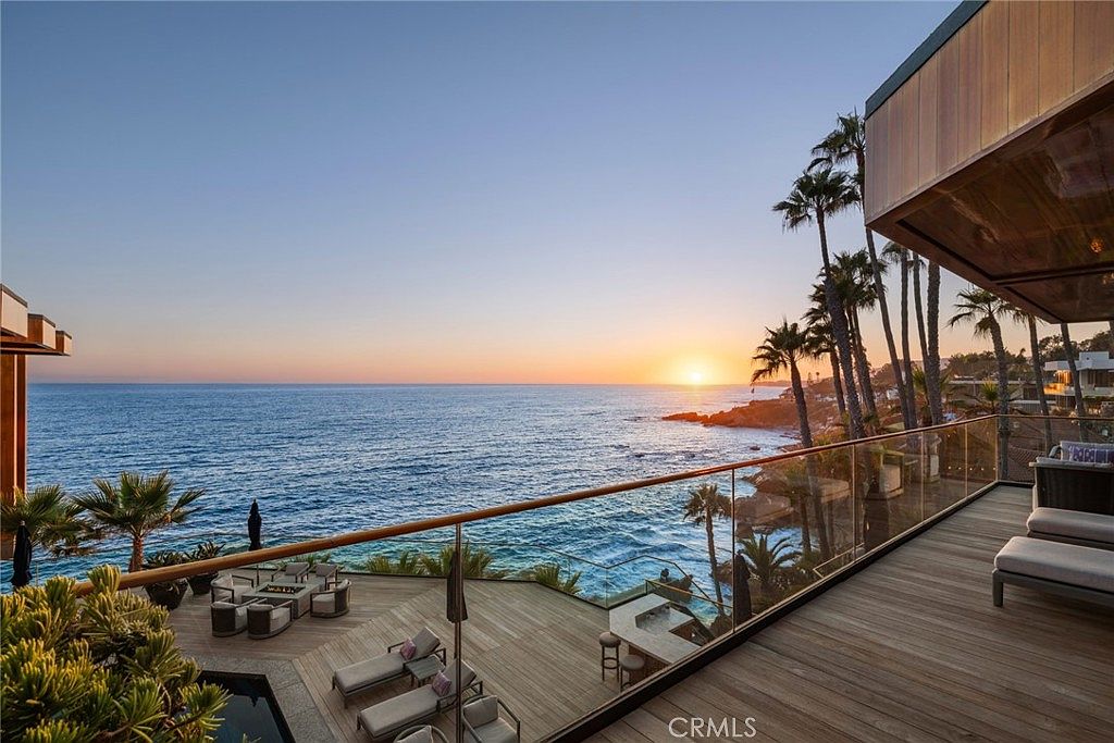 This stunning elevated balcony offers a panoramic, unobstructed view of the ocean at sunset, featuring a sleek glass railing that maintains the visual connection to the horizon. Below, a multi-level wooden deck includes a lounge area with comfortable seating and a fire pit, seamlessly integrating with the coastal landscape. The perspective captures the luxurious outdoor living space from an upper-level vantage point, highlighting the serene atmosphere and prime waterfront location.