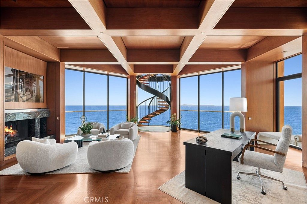 This expansive, light-filled room serves as a sophisticated home office and lounge area, featuring warm wood-paneled walls and a coffered ceiling. The space is defined by large windows that provide panoramic ocean views, a central spiral staircase, and a dark wood desk positioned to overlook the water. A cozy seating area with four plush chairs and a fireplace with a dark stone surround completes the luxurious, tranquil atmosphere.