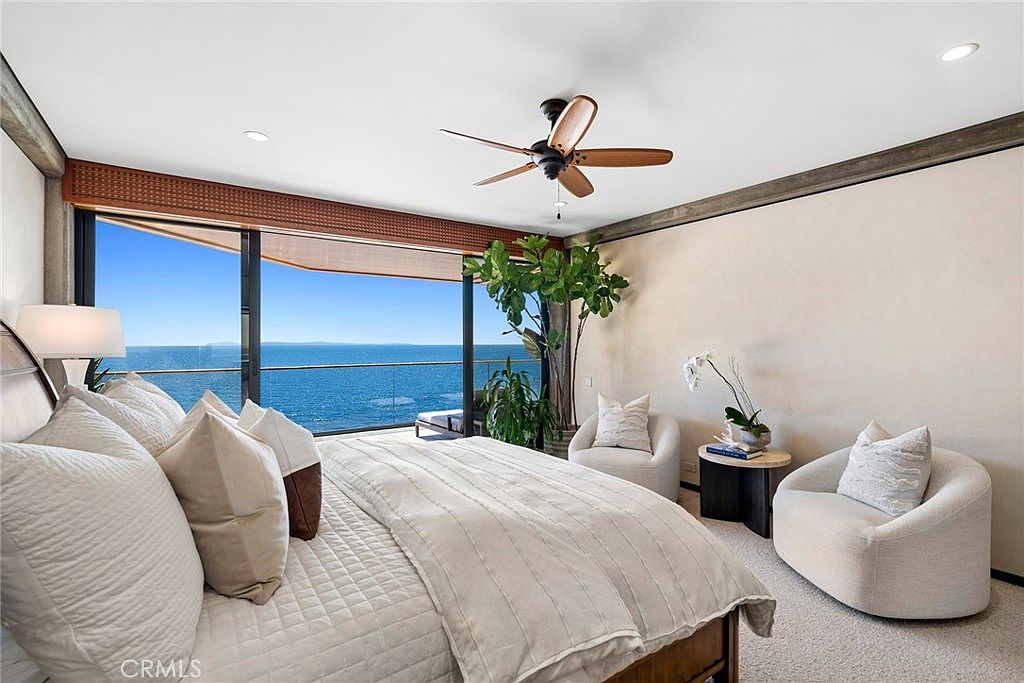 This serene primary bedroom features a neutral color palette and a large bed positioned to face floor-to-ceiling windows that offer an expansive, unobstructed view of the ocean. The room includes two comfortable, rounded accent chairs and a small side table, creating a cozy seating area. A ceiling fan hangs above, and a large potted plant adds a touch of greenery to the bright, airy space.