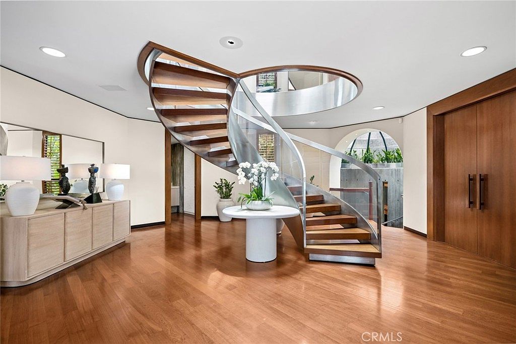 This grand entryway features a striking, sculptural spiral staircase with wooden treads and a curved metal railing as its central focal point. A circular table with a floral arrangement sits beneath the staircase, while a long console table with lamps lines the left wall. The space is finished with warm wood flooring and clean, neutral walls, creating an elegant and sophisticated atmosphere.