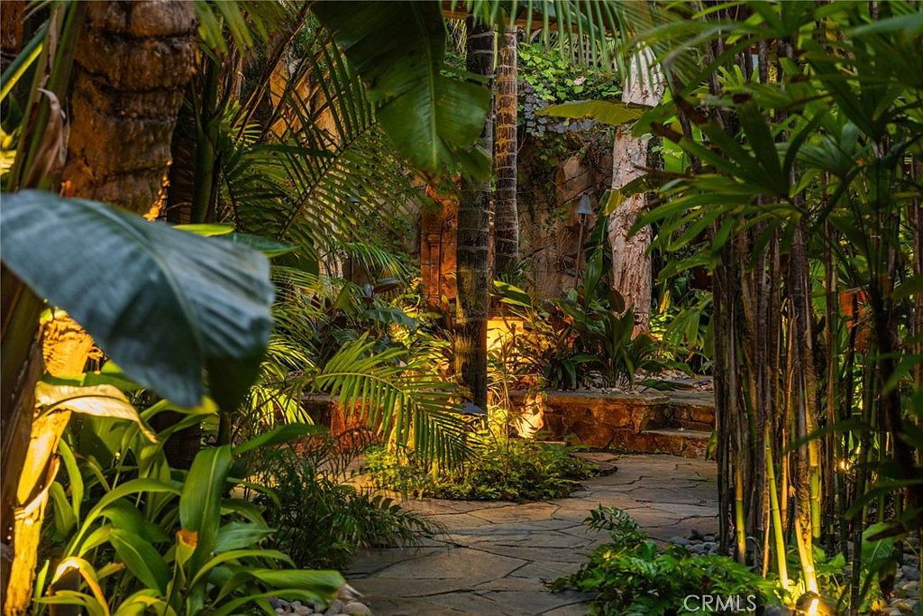 This lush, tropical-inspired garden path features irregular flagstone paving winding through dense foliage, including palm trees and various ornamental plants. Warm landscape lighting illuminates the path and the textured stone wall in the background, creating an inviting and serene atmosphere. The perspective is at eye level, drawing the viewer into the tranquil, secluded outdoor space.