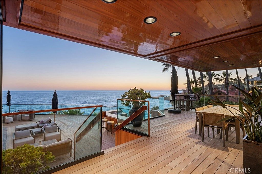 This expansive, multi-level wooden deck offers a luxurious outdoor living space with panoramic ocean views. The area features a comfortable lounge seating arrangement on the lower level and a dining set on the upper deck, all sheltered by a polished wood-paneled ceiling with recessed lighting. Glass railings provide an unobstructed view of the coastline, creating a seamless transition between the home and the horizon.