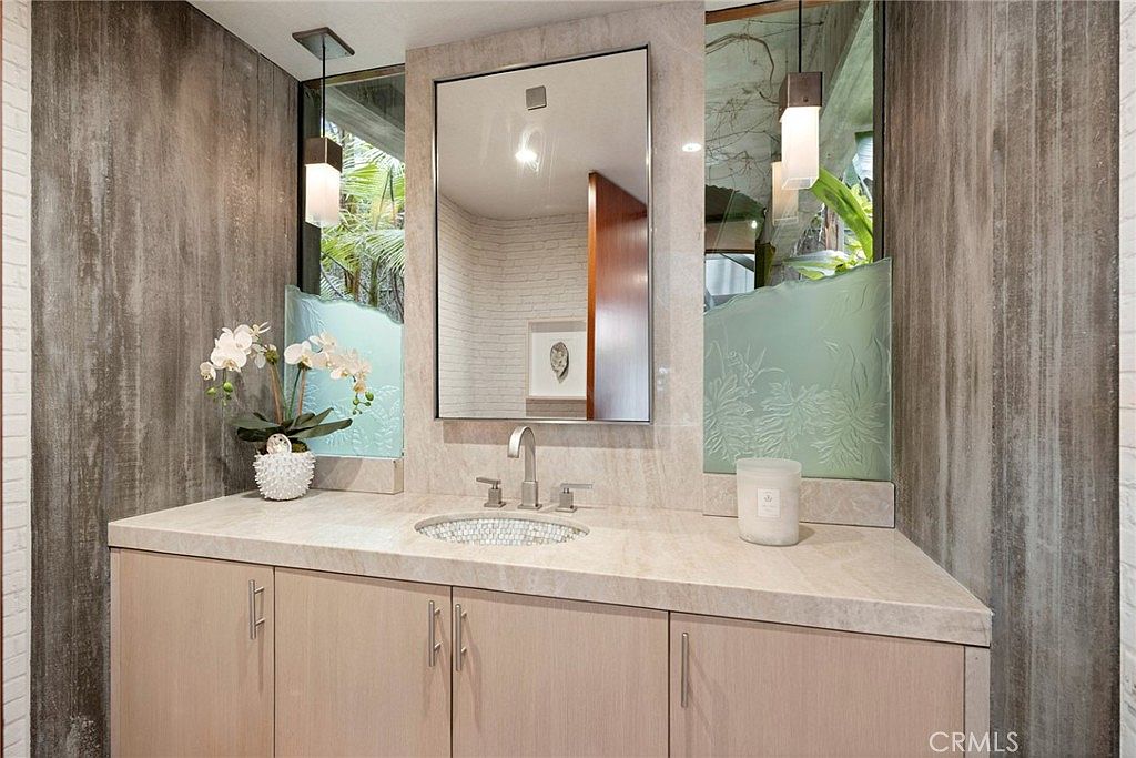 This bathroom vanity area features a light-toned wood cabinet topped with a neutral stone countertop and a mosaic-tiled sink basin. A rectangular mirror is centered between two frosted glass panels, which are illuminated by modern pendant lights hanging from above. The walls are finished with textured, vertical-patterned panels, creating a sophisticated and contemporary aesthetic.