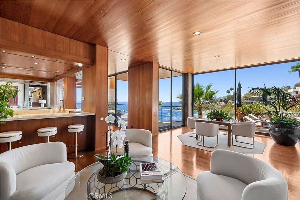 This sophisticated living area features warm wood-paneled ceilings and walls, creating a seamless, high-end aesthetic. The space is anchored by a circular glass coffee table surrounded by plush white armchairs, with a dining area positioned near large windows that offer expansive ocean views. The open-concept layout includes a built-in bar area, emphasizing a luxurious, coastal-modern design perfect for entertaining.