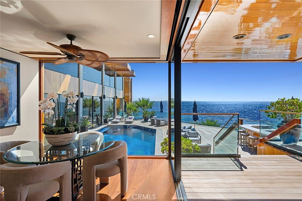 This sophisticated dining area features a glass-topped table surrounded by modern, neutral-toned chairs, positioned directly beside expansive, floor-to-ceiling glass walls that slide open to a deck. The space offers a seamless transition to an outdoor pool area and an unobstructed, panoramic view of the ocean. A wooden ceiling with a recessed fan adds warmth to the contemporary design, creating an airy and luxurious atmosphere.