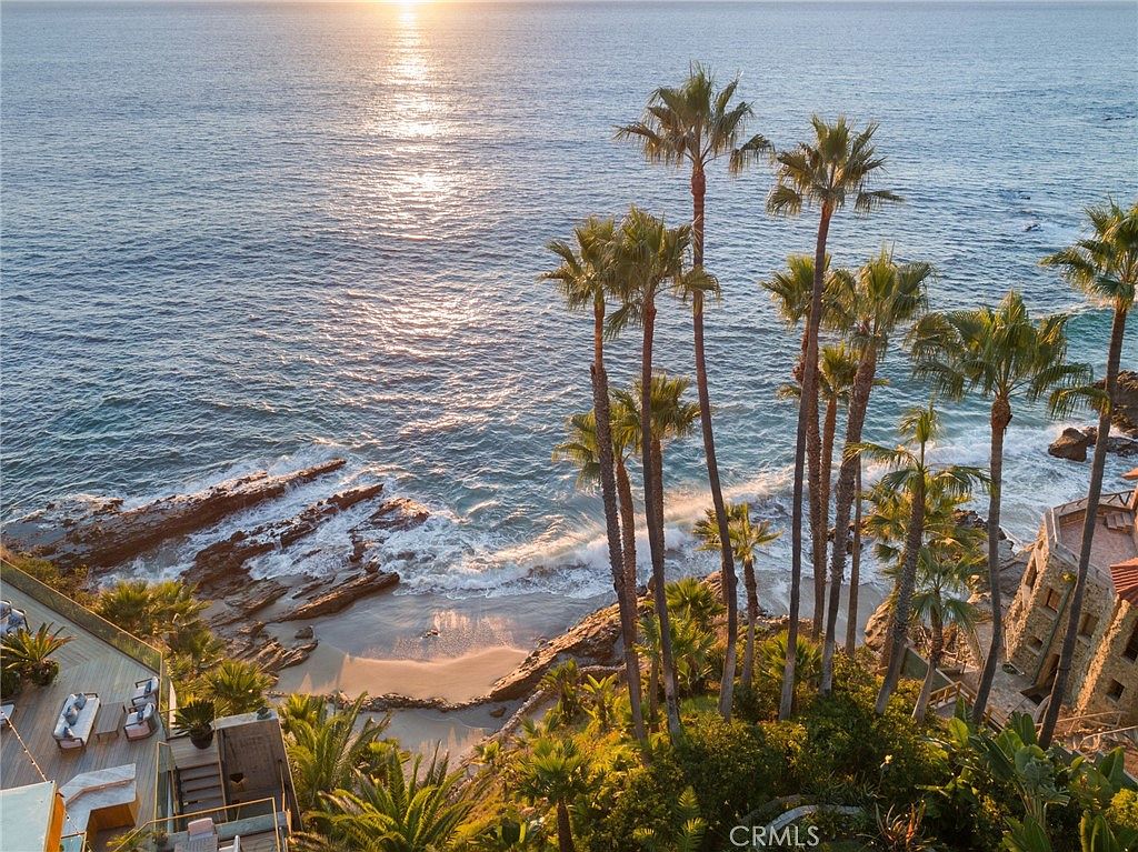 This high-angle aerial view captures a stunning coastal property overlooking a secluded beach and the Pacific Ocean at sunset. The scene features a lush landscape filled with tall palm trees, rugged rock formations along the shoreline, and a multi-level deck area with outdoor seating. The warm, golden light reflecting off the water creates a serene and luxurious atmosphere, highlighting the property's prime oceanfront location.