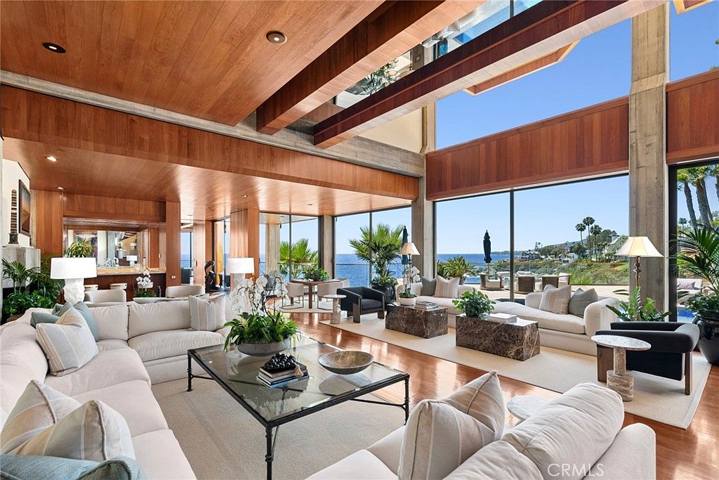 This expansive, open-concept living room features high ceilings with exposed structural beams and warm wood paneling. Large glass walls provide panoramic ocean views, seamlessly connecting the interior space to the outdoor patio. The room is furnished with multiple plush white sofas, stone-topped coffee tables, and elegant lighting, creating a sophisticated and airy atmosphere.