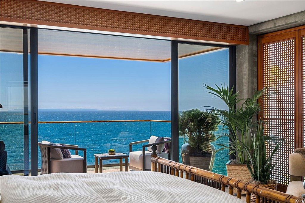 This primary bedroom features a direct, unobstructed view of the ocean through expansive floor-to-ceiling glass sliding doors that open onto a private balcony. The room is furnished with a comfortable bed in the foreground and a seating area on the balcony, creating a serene and luxurious atmosphere. A large potted plant adds a touch of greenery to the interior, while the warm wood-toned ceiling detail and textured wall accents enhance the sophisticated coastal aesthetic.