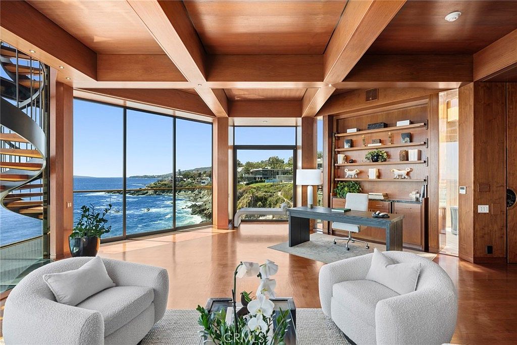 This sophisticated home office features a warm, wood-paneled aesthetic with a coffered ceiling and expansive floor-to-ceiling windows that frame a stunning ocean view. The space is furnished with a modern desk, a pair of plush white armchairs, and built-in shelving units that provide ample storage and display space. The room is bathed in natural light, creating a serene and professional atmosphere that seamlessly blends indoor comfort with the coastal landscape.