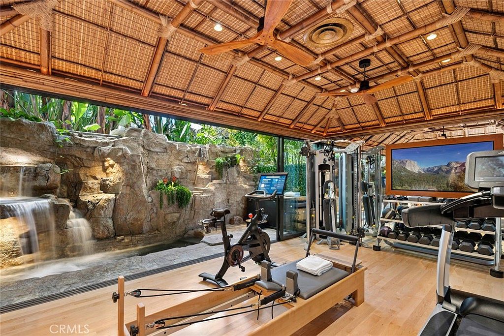 This unique home gym features a tropical-inspired aesthetic with a vaulted bamboo-style ceiling and a dramatic indoor rock wall with a built-in waterfall feature. The space is equipped with a stationary bike, a Pilates reformer, and a multi-station weight machine, all set upon light wood flooring. Large glass panels provide an open view of the lush greenery outside, creating a serene and immersive workout environment.