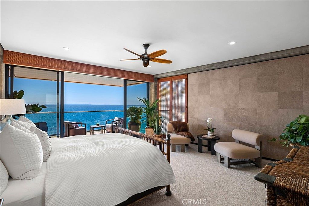 This primary bedroom features a serene, coastal-inspired aesthetic with a direct view of the ocean through expansive glass sliding doors. The room is furnished with a plush bed, a pair of accent chairs, and a small side table, all set against a textured, neutral-toned wall. A ceiling fan hangs above, and the space is bathed in natural light, creating a tranquil and luxurious atmosphere.
