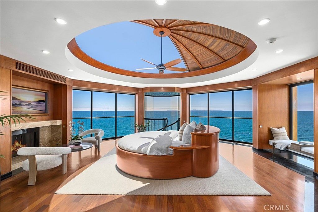 This luxurious primary bedroom features a unique circular layout with a central round bed positioned beneath a dramatic domed ceiling with a fan. The room is surrounded by expansive glass walls that provide panoramic ocean views, creating a seamless connection to the horizon. A fireplace with a stone surround adds warmth to the space, while the warm wood paneling and polished wood floors enhance the sophisticated, coastal-inspired aesthetic.