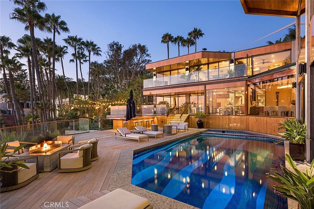 This luxurious multi-level residence features a striking modern design with extensive glass walls and warm wood-toned siding. The expansive outdoor deck includes a custom-tiled swimming pool with a unique wave pattern, a cozy fire pit lounge area, and a built-in bar, all set against a backdrop of lush palm trees. The scene is captured at dusk, with warm string lights and interior illumination creating an inviting and sophisticated atmosphere.