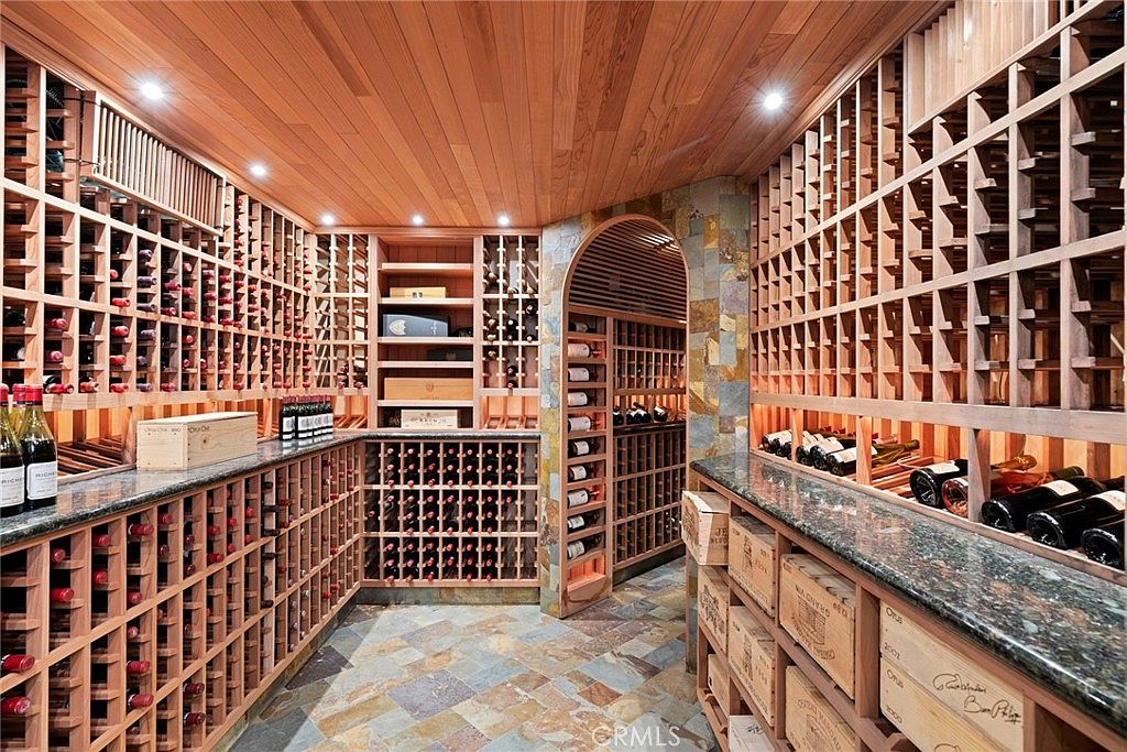 This custom wine cellar features extensive wooden racking systems that span the walls, providing ample storage for a large collection. The room is accented by a stone-clad arched entryway and a multi-colored stone tile floor, creating a sophisticated and rustic atmosphere. Granite countertops provide a functional surface for display and service, while recessed lighting highlights the warm wood tones throughout the space.