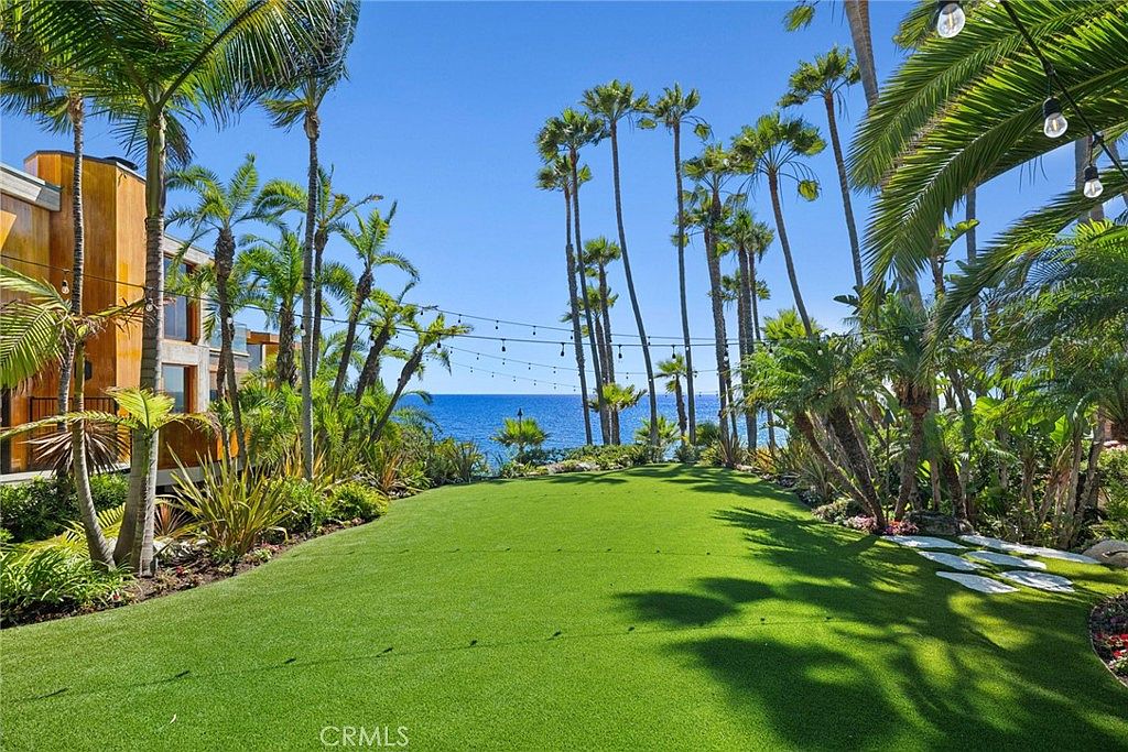 This expansive, manicured lawn serves as a lush backyard oasis, framed by a variety of mature palm trees and tropical landscaping. String lights are suspended across the yard, creating an inviting atmosphere that leads the eye toward a stunning, unobstructed view of the ocean horizon. The scene is captured from a ground-level perspective, emphasizing the seamless transition between the private green space and the coastal scenery.
