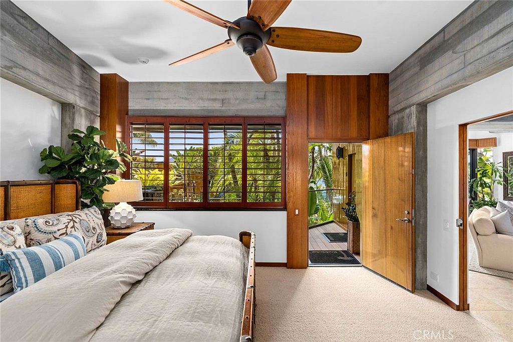 This primary bedroom features a warm, tropical-inspired aesthetic with rich wood paneling and textured concrete walls. A large bed with patterned pillows sits beneath a ceiling fan, while a set of louvered shutters allows natural light to filter in from the outside. An open doorway leads to a private balcony, and a glimpse into an adjacent living area is visible through a secondary doorway.