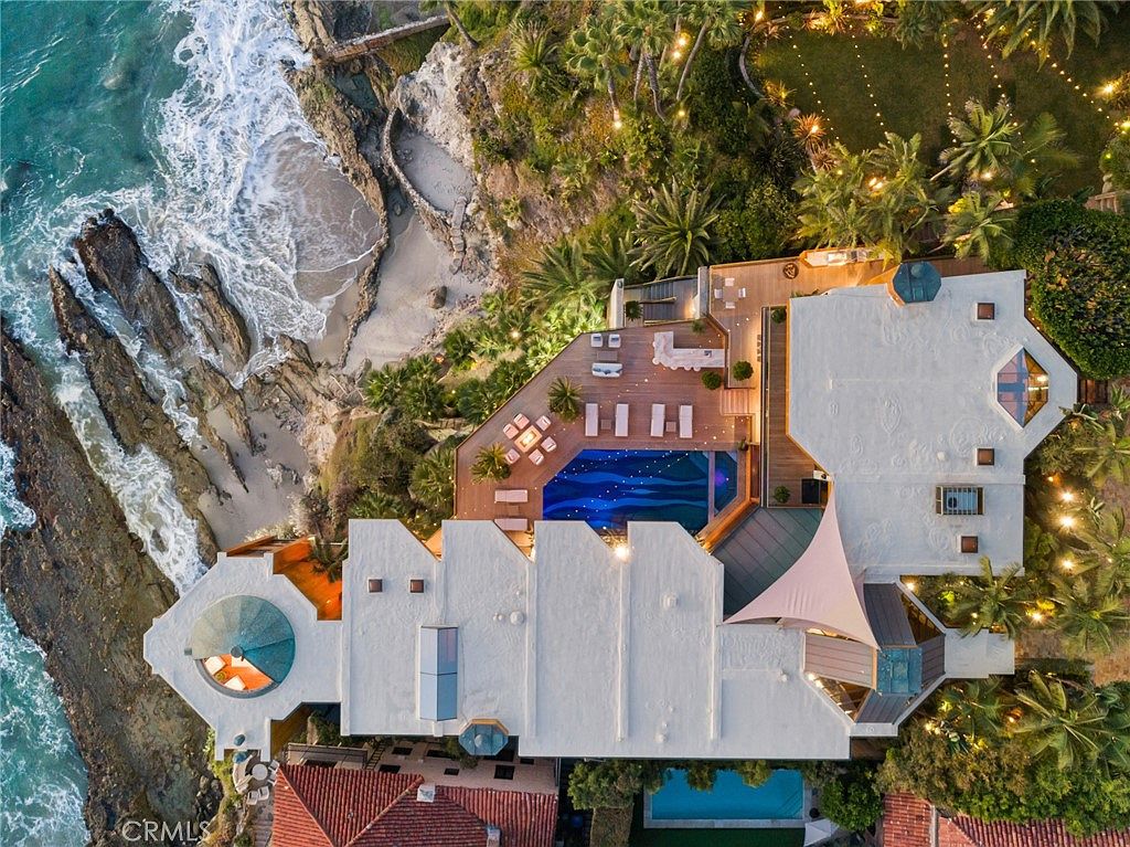 This dramatic aerial perspective captures a luxury coastal estate perched directly above the ocean, showcasing its unique architectural design and expansive outdoor living spaces. The property features a custom-shaped swimming pool, multiple tiered wooden decks, and a private staircase leading down to a secluded sandy cove. Lush landscaping, string lighting, and a distinctive domed roof structure highlight the high-end, resort-like atmosphere of this cliffside residence.