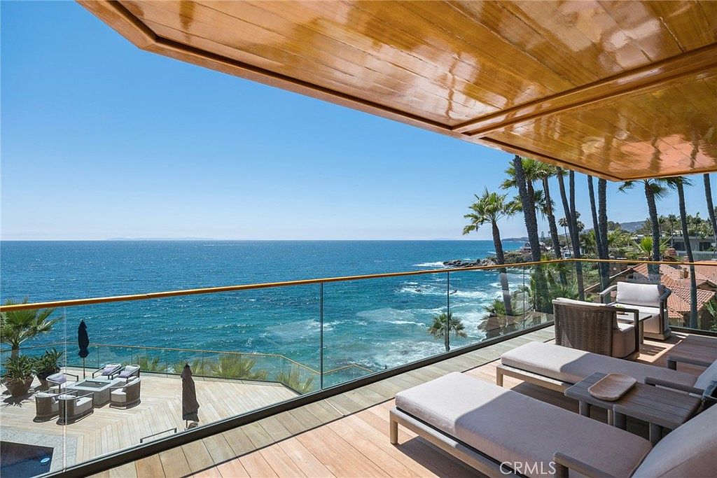 This expansive, elevated balcony offers a panoramic, unobstructed view of the ocean and coastline. The space is furnished with modern lounge chairs and features a sleek glass railing that maintains the visual connection to the sea, while a polished wood ceiling adds a warm, sophisticated touch. Below, a lower deck area with outdoor seating and a fire pit provides additional space for relaxation, creating a seamless transition between indoor luxury and the natural coastal environment.