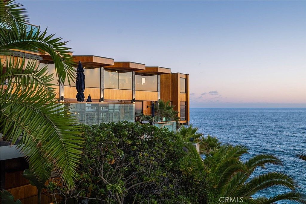 This striking modern residence is perched on a coastal cliff, showcasing a multi-level architectural design with warm wood siding and expansive glass walls. The structure features deep, cantilevered rooflines and integrated balconies that offer unobstructed views of the ocean at sunset. Lush tropical foliage in the foreground frames the home, emphasizing its secluded and luxurious cliffside setting.