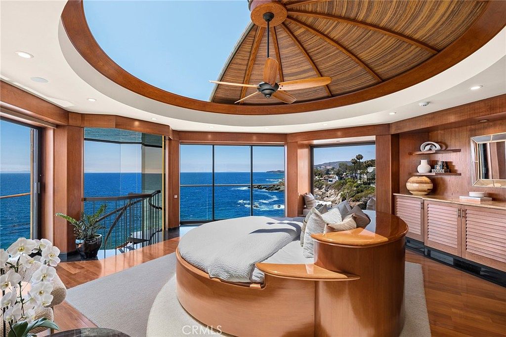 This luxurious primary bedroom features a unique circular layout with a central round bed positioned under a striking domed ceiling with a wooden fan. The room is surrounded by expansive glass walls that offer panoramic views of the ocean and coastline. Warm wood paneling, built-in shelving, and polished floors create a sophisticated, high-end atmosphere.