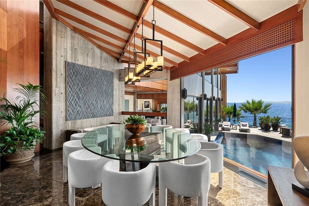 This sophisticated dining room features a large glass-topped table surrounded by white upholstered chairs, positioned under a modern geometric chandelier. The space is defined by a textured concrete wall and exposed wood ceiling beams, creating a blend of industrial and organic aesthetics. Large glass walls provide a seamless transition to an outdoor pool area with expansive ocean views, emphasizing a luxurious indoor-outdoor living experience.