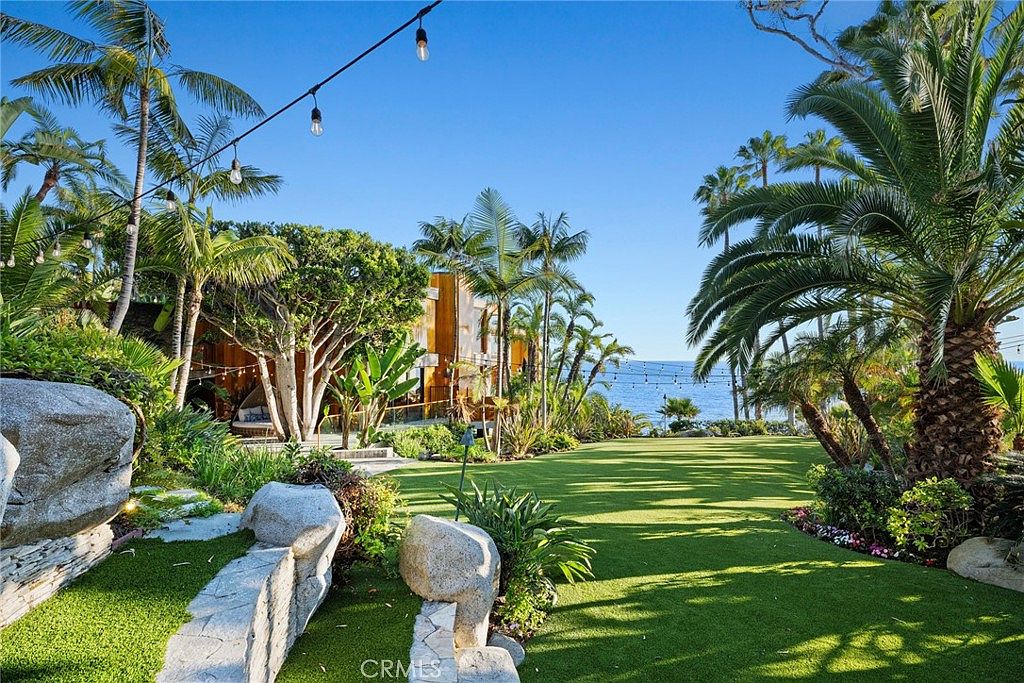 This expansive, manicured lawn overlooks the ocean, framed by lush tropical landscaping and mature palm trees. A string of overhead lights creates an inviting atmosphere, leading the eye toward the modern residence nestled among the greenery. The scene captures a serene, coastal lifestyle with a seamless transition between the private garden and the natural beauty of the sea.