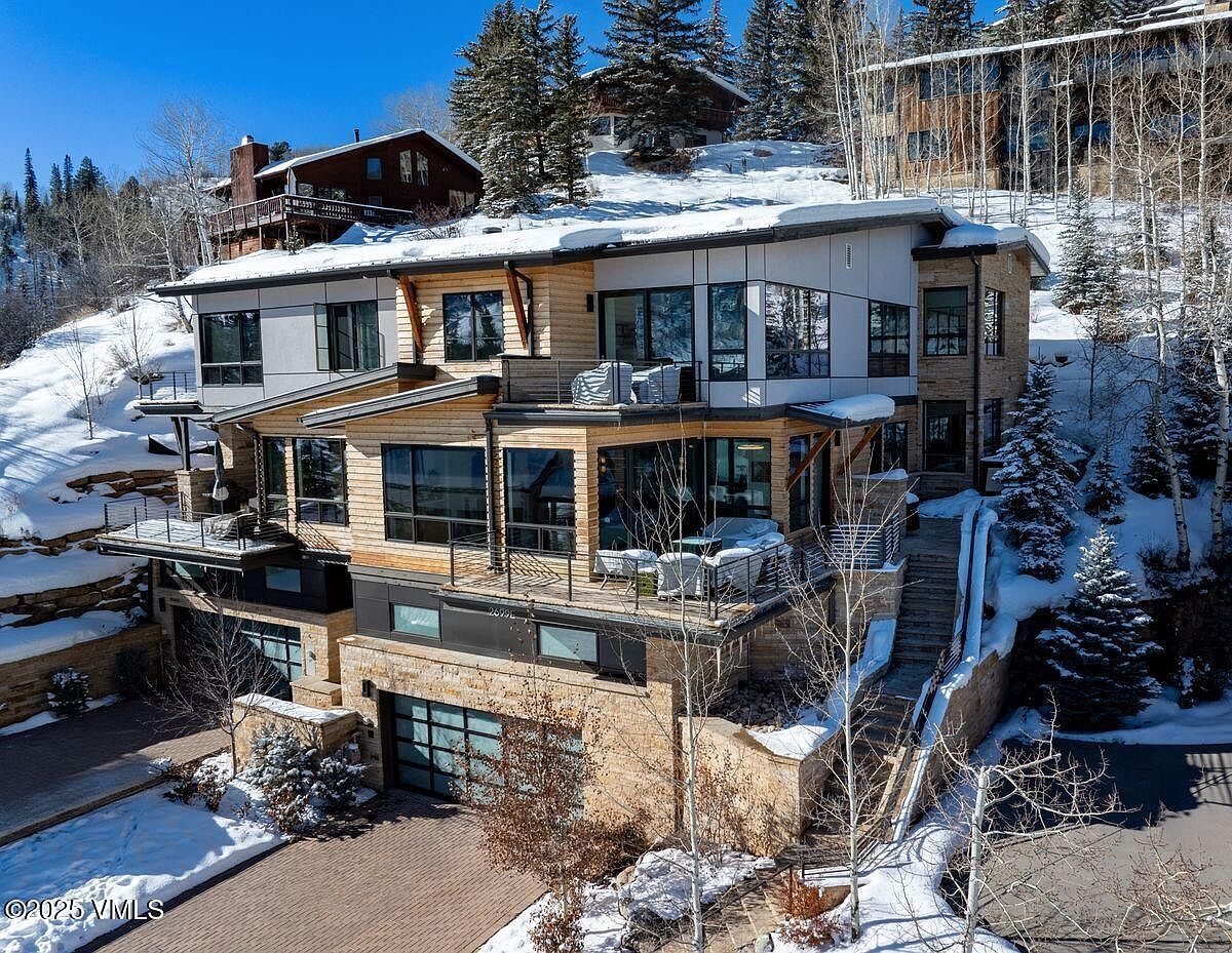 This is a front exterior view of a modern multi-level home nestled on a snowy hillside. The house features a combination of stone, wood, and metal siding, with large windows and multiple balconies offering stunning views. A paved driveway leads to a garage, and stairs provide access to the upper levels, creating an inviting and luxurious mountain retreat.