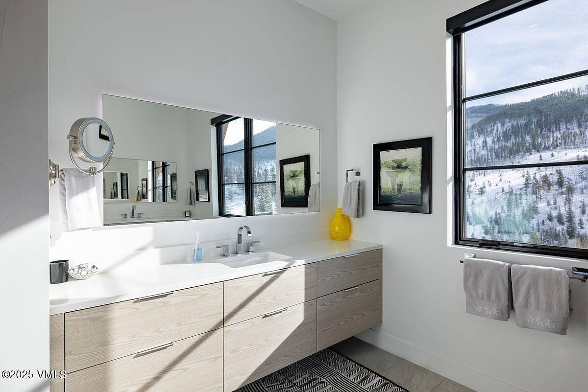 This is a modern bathroom featuring a floating vanity with light wood-grain drawers and a white countertop. A large mirror reflects the window view, enhancing the natural light. The room is decorated with minimalist artwork and accessories, creating a clean and serene atmosphere.