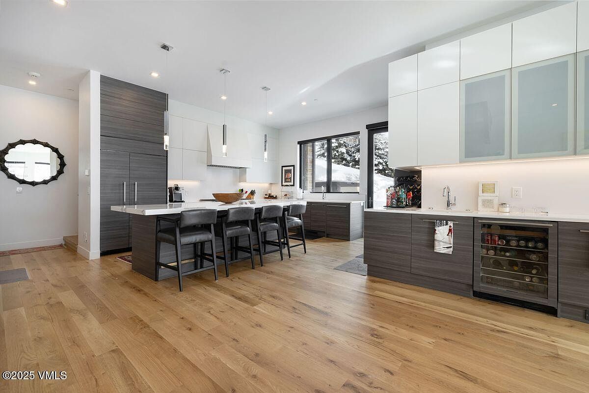 This is a modern kitchen with a large island featuring seating and a white countertop. The cabinetry is a mix of white and gray wood grain, with stainless steel appliances and a wine fridge. The hardwood floors add warmth to the space, and the large windows provide natural light and a view of the snowy outdoors.