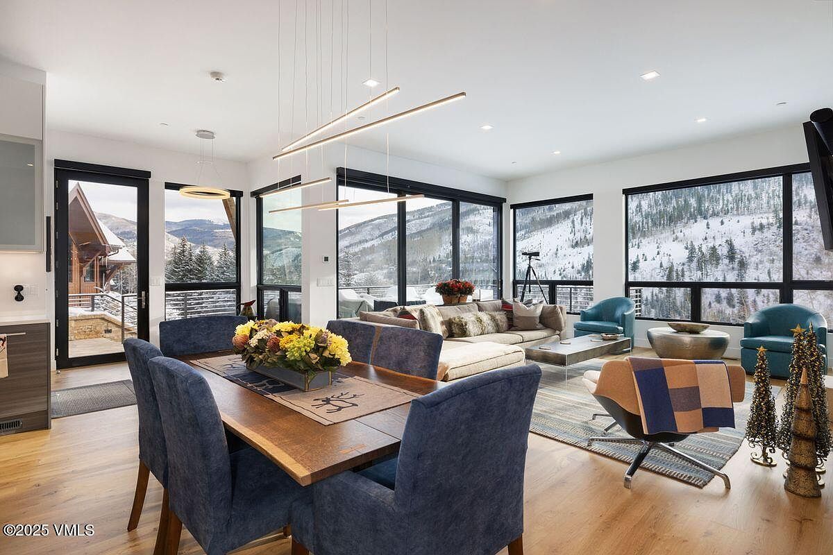 This is an interior shot of a modern living and dining area with an open floor plan. The dining area features a wooden table with blue upholstered chairs and a floral centerpiece, while the living area includes a comfortable sofa, armchairs, and a view of a snow-covered mountain through large windows. The room is well-lit with natural light and modern lighting fixtures, creating a warm and inviting atmosphere.
