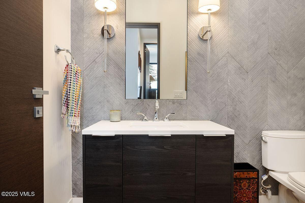 This is a well-lit bathroom featuring a modern vanity with a dark wood finish and a white countertop. The walls are adorned with a stylish gray herringbone tile pattern, complemented by sleek sconce lighting on either side of the mirror. A colorful towel hangs neatly on a rack, adding a touch of vibrancy to the space.