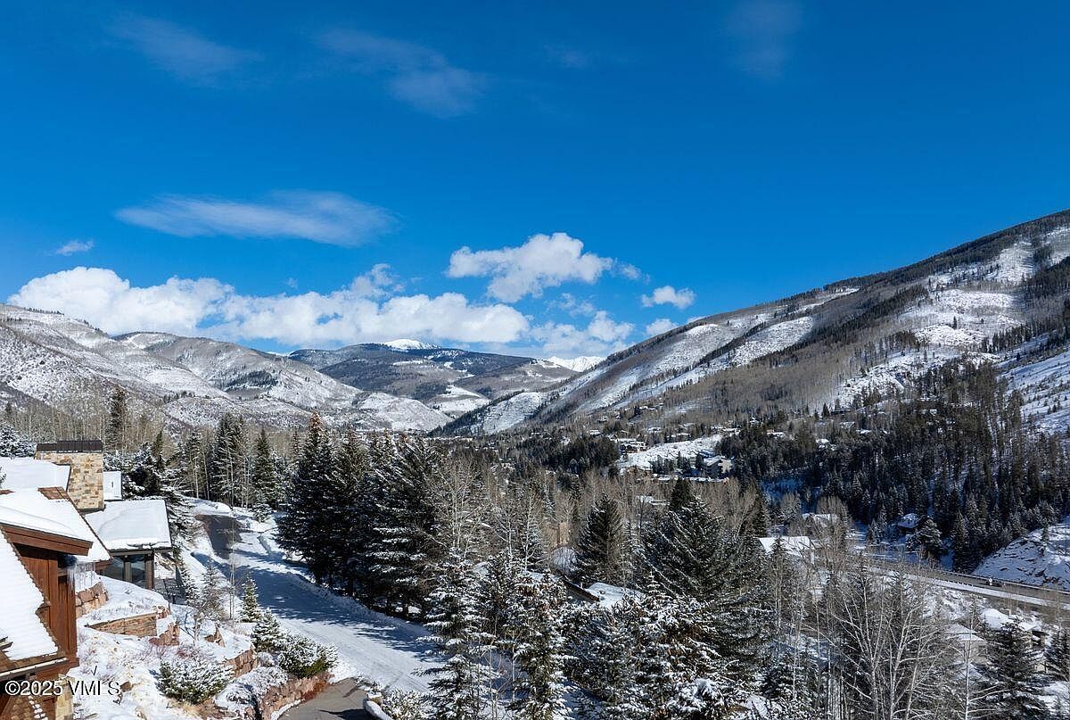 This image showcases a picturesque mountain landscape with snow-covered peaks and evergreen trees. A residential property is nestled among the trees, featuring a snow-covered roof and a glimpse of its exterior. The clear blue sky and bright sunlight create a serene and inviting atmosphere, highlighting the property's scenic location.