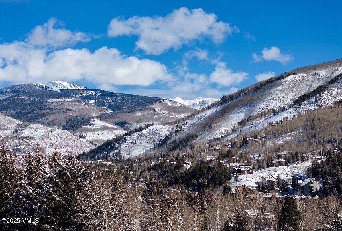 This aerial view showcases a picturesque mountain landscape with snow-dusted peaks and slopes. Nestled among the trees are residential homes, suggesting a serene and secluded community. The clear blue sky with scattered clouds enhances the scenic beauty, making it an appealing location for real estate.