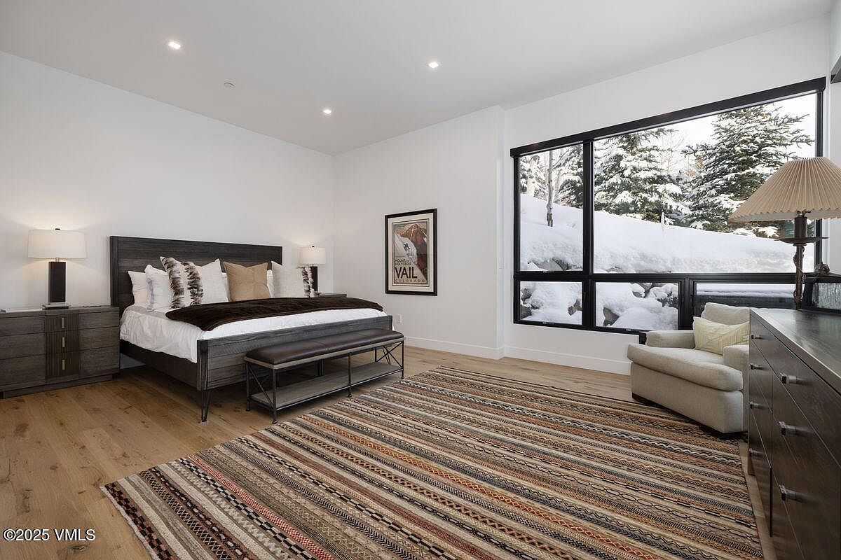 This is a primary bedroom featuring a large window with a view of a snowy landscape. The room is decorated in neutral tones with a dark wood bed frame and matching nightstands. A striped rug adds texture to the light wood flooring, and an armchair sits near the window, creating a cozy reading nook.