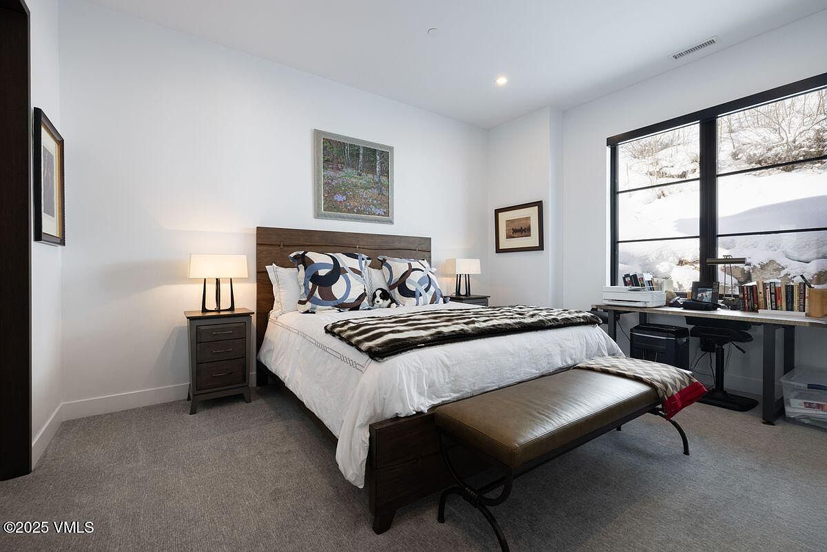 This is a primary bedroom featuring a wooden bed frame with decorative pillows and a striped throw blanket. The room has a neutral color palette with white walls and gray carpeting. A desk area with a window provides natural light, and the overall impression is cozy and inviting.