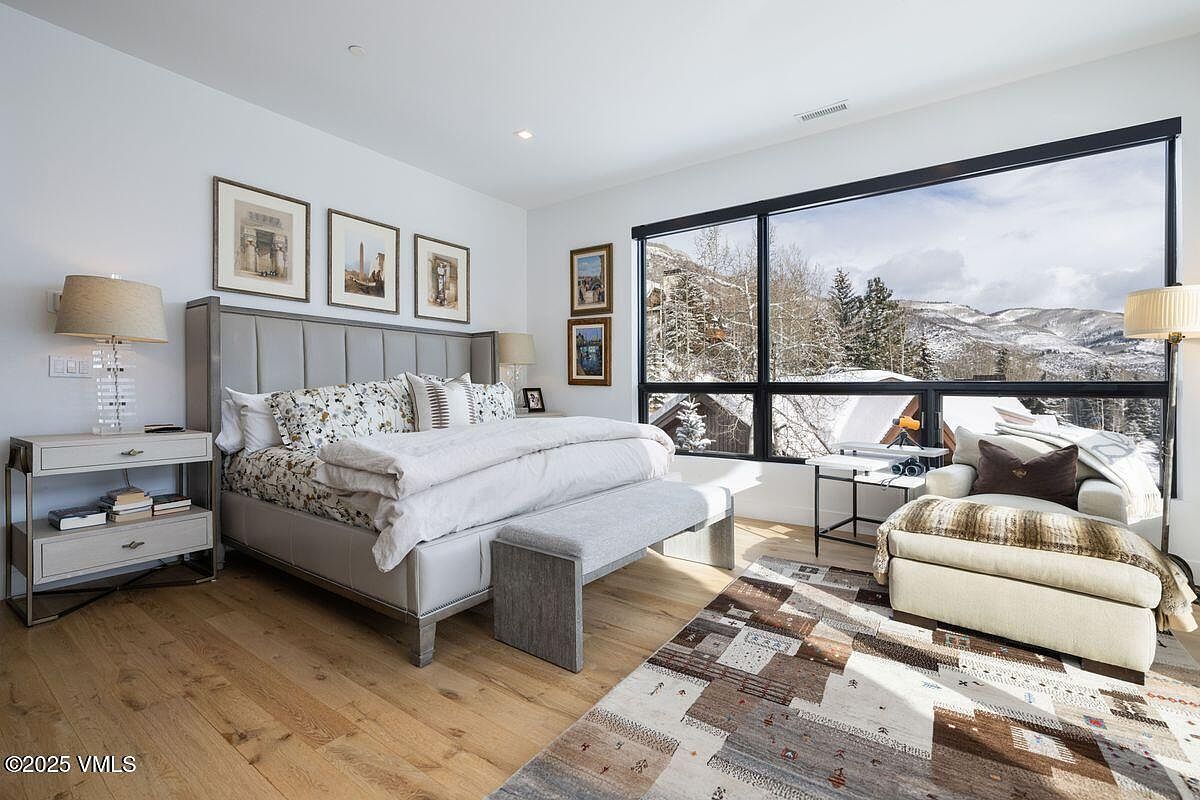 This primary bedroom features a large window with mountain views, a king-sized bed with a padded headboard, and a neutral color palette. The room is well-lit and includes a side table with a lamp, artwork on the walls, and a cozy seating area with a patterned rug, creating a serene and inviting atmosphere. The perspective is from the doorway, showcasing the entire room.