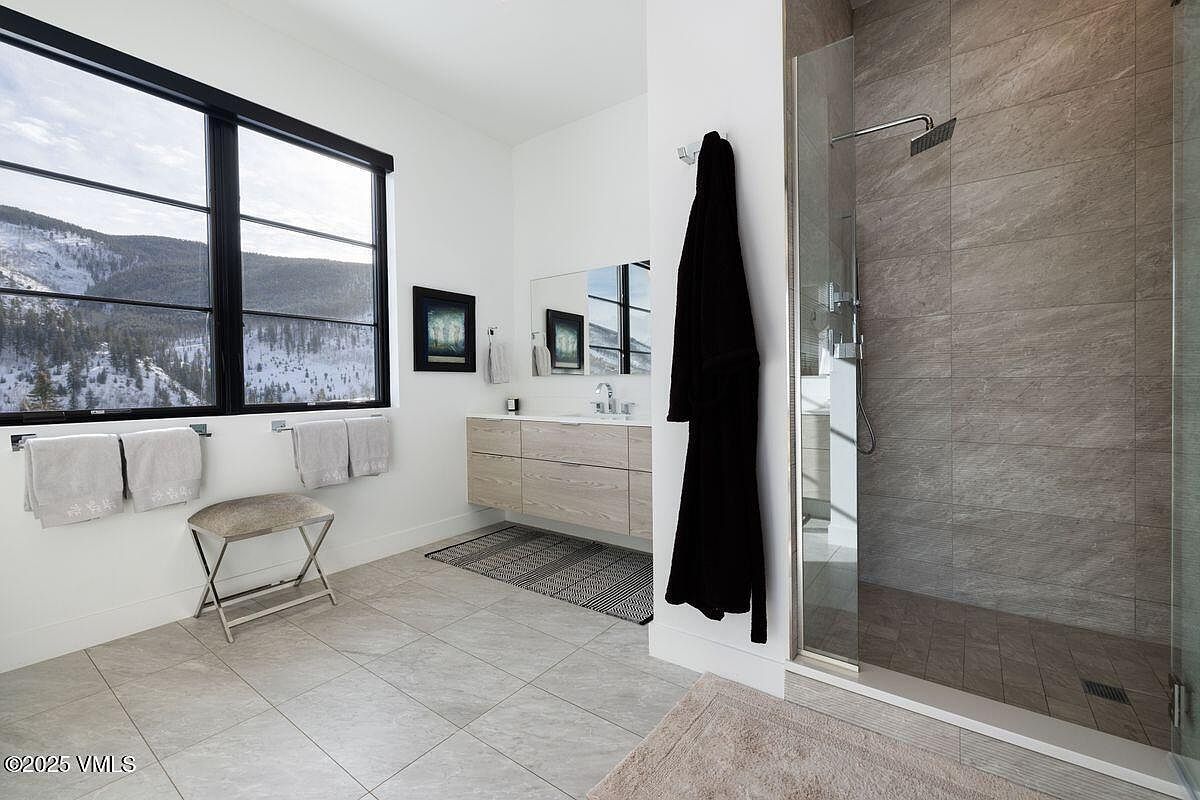 This is a modern primary bathroom featuring a large window with mountain views, a floating vanity with light wood drawers, and a glass-enclosed shower with gray tile. A black robe hangs near the shower, adding a touch of luxury, while neutral-toned towels and a bench provide comfort. The overall impression is clean, bright, and spa-like.