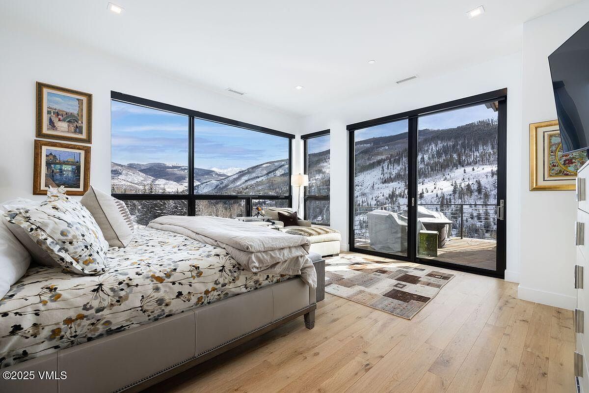 This is a bright and airy primary bedroom featuring large windows with stunning mountain views. The room is furnished with a bed with floral patterned bedding, a chaise lounge, and light wood flooring. Artwork adorns the walls, adding a touch of elegance to the space.
