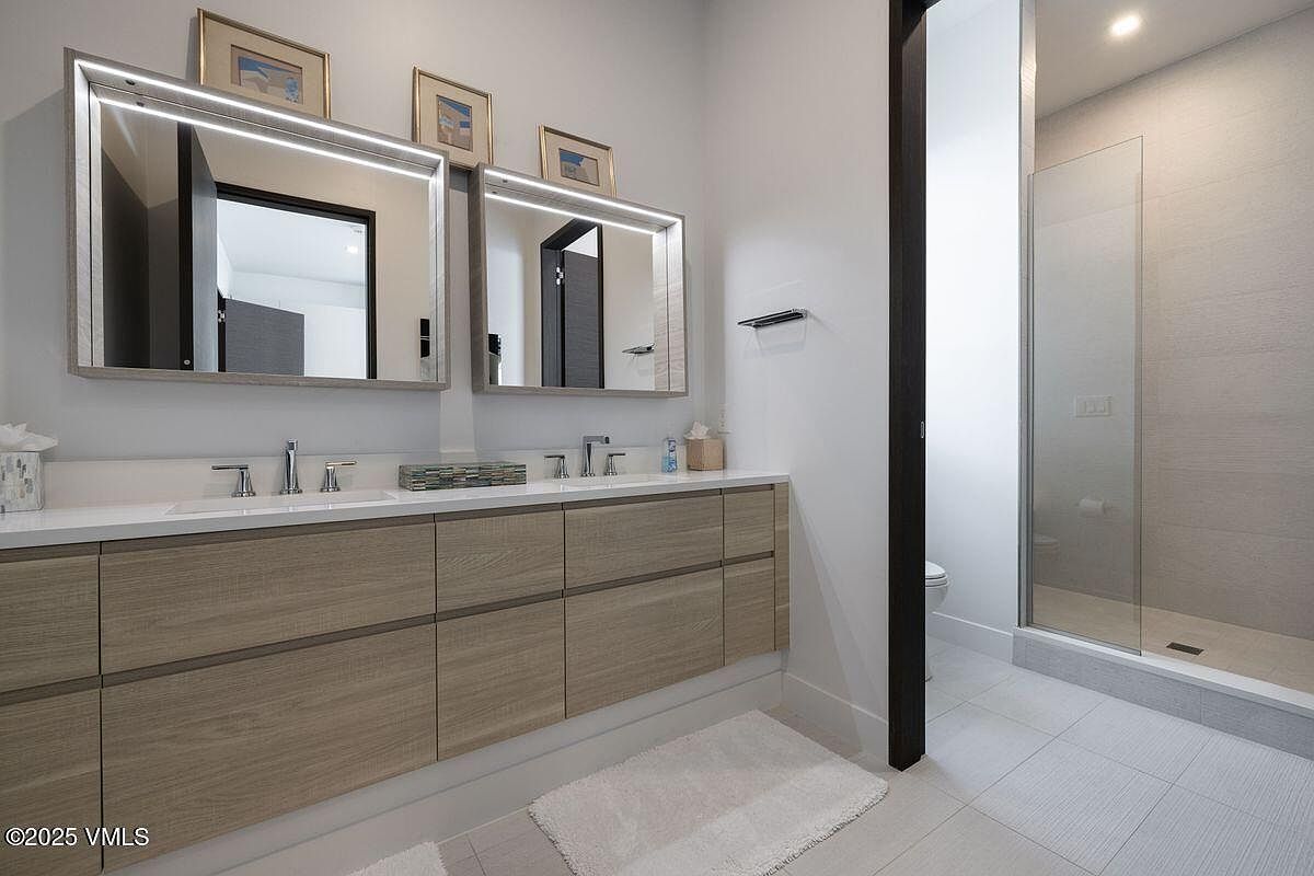 This is a modern bathroom featuring a double vanity with light wood cabinetry and a white countertop. Two illuminated mirrors hang above the sinks, and framed artwork is displayed above them. The bathroom includes a glass-enclosed shower and light gray tile flooring, creating a clean and contemporary aesthetic.