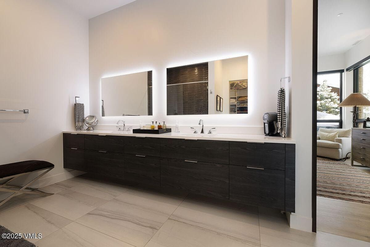 This is a modern primary bathroom featuring a double vanity with dark wood cabinetry and a white countertop. Two large, illuminated mirrors hang above the sinks. The flooring consists of large, light-colored tiles with subtle veining, and a glimpse into an adjacent room reveals a cozy seating area, suggesting a spacious and luxurious master suite.