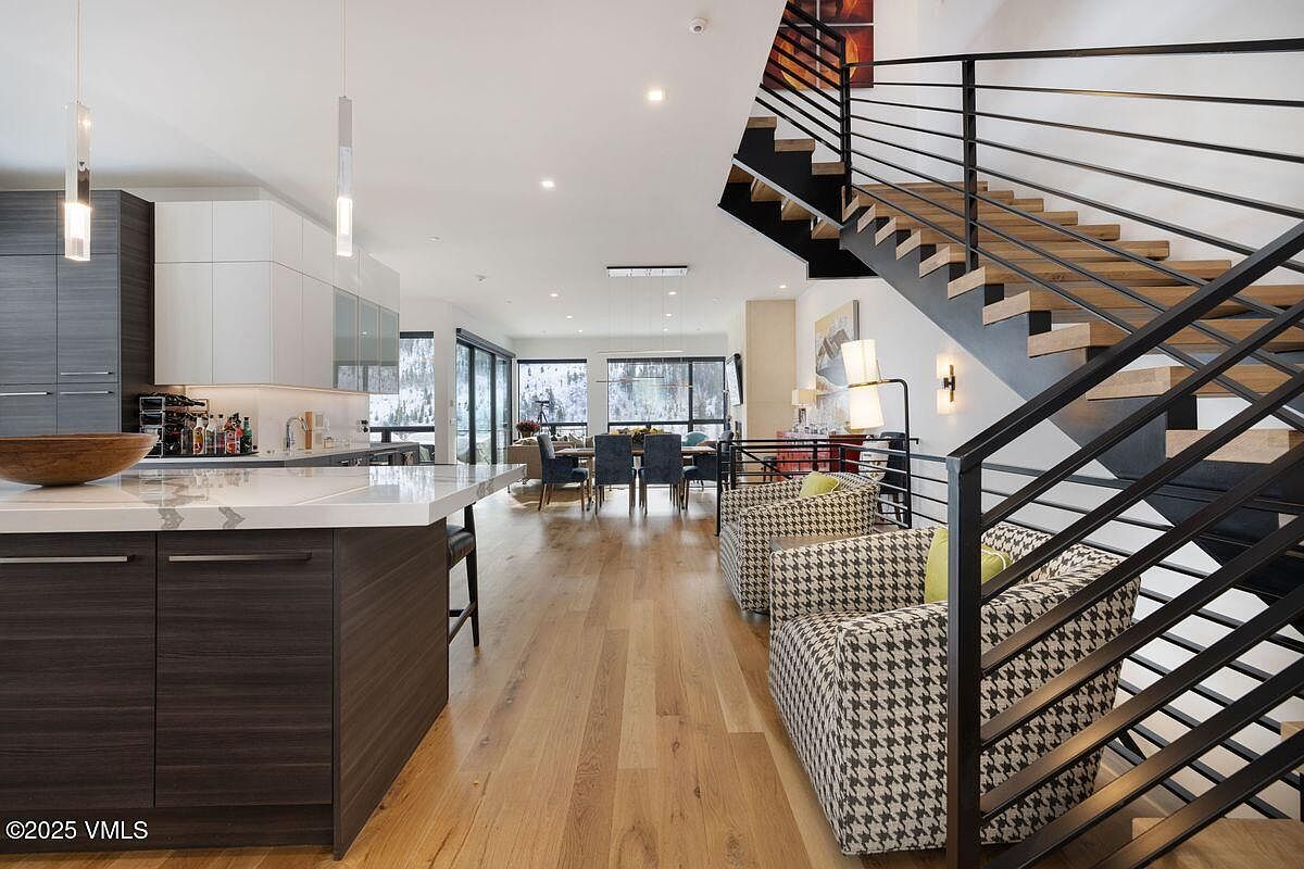 This interior shot showcases a modern living space with an open floor plan, integrating the kitchen, dining area, and living room. The design features a striking staircase with black metal railings and wooden steps, complemented by houndstooth patterned armchairs. Large windows provide ample natural light and views of the outdoors, enhancing the spacious and airy feel of the home.