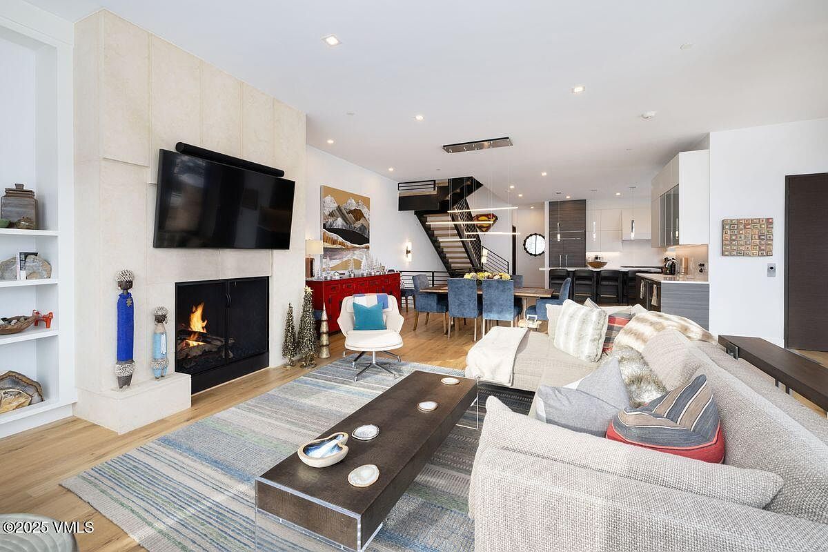 This is an interior shot of a modern living room featuring a fireplace with a mounted TV above it, a comfortable sofa, and a stylish coffee table. The open floor plan seamlessly connects the living area to the dining space and kitchen, creating a spacious and inviting atmosphere. The room is well-lit with recessed lighting and natural light, highlighting the contemporary design and high-end finishes.