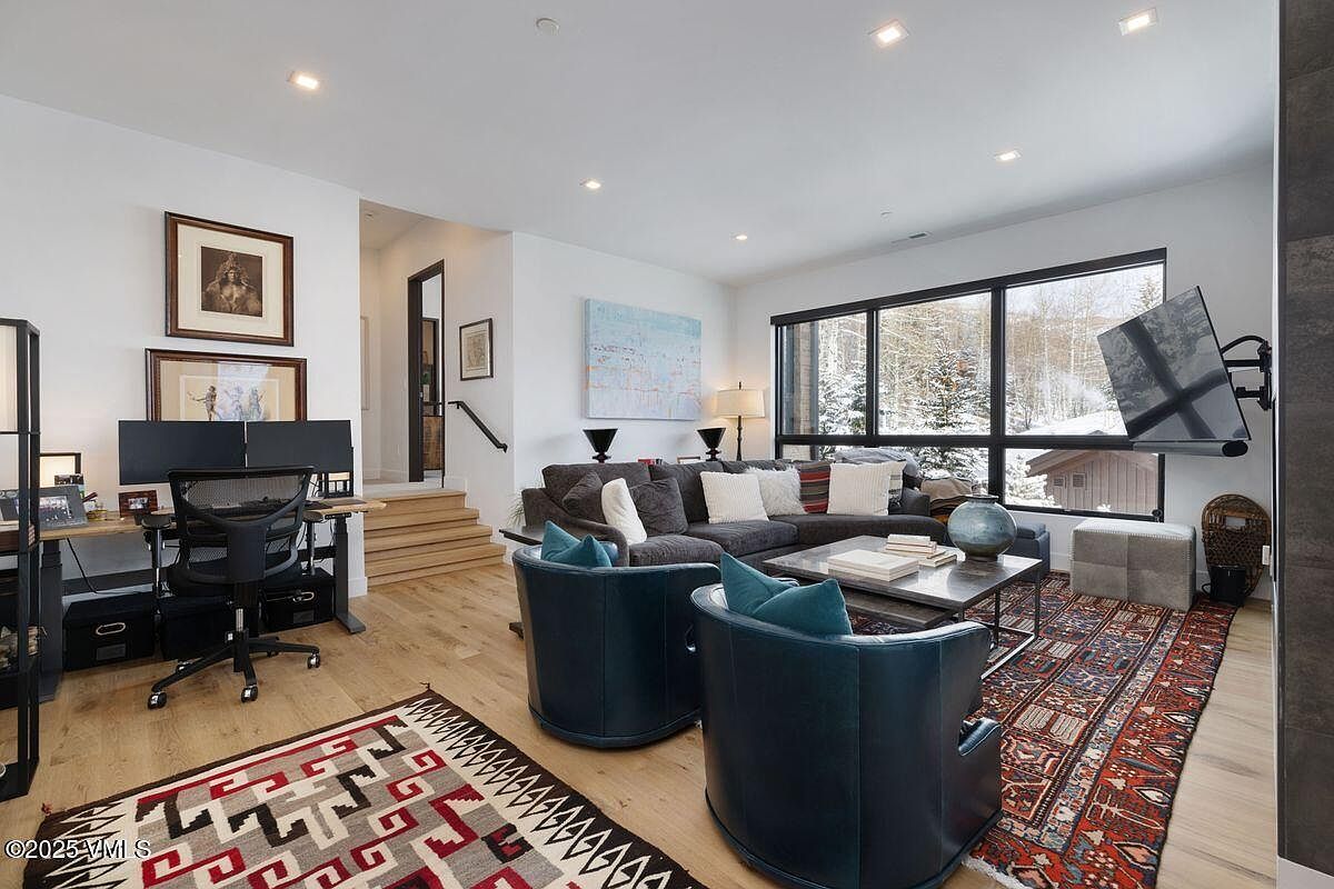 This is an interior shot of a living room featuring a comfortable gray sectional sofa adorned with decorative pillows, complemented by two teal swivel chairs. A modern coffee table sits atop a patterned rug, and a large window offers a view of a snow-covered landscape. The room also includes a dedicated office space with a desk and monitors, creating a versatile and inviting atmosphere.